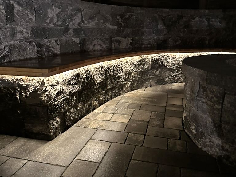 Geschwungene Sitzmauer aus Naturstein mit Holzauflage und LED-Licht am Abend